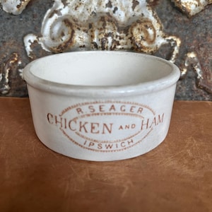 May include: A small, round, off-white ceramic pot with a flat rim. The front features a brown oval stamp with the text R. SEAGER, CHICKEN AND HAM, and IPSWICH printed in a clear, sans-serif font. The surface has a smooth, matte finish.