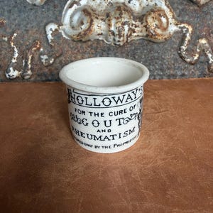 May include: A small, cylindrical white ceramic ointment pot featuring black printed text that reads: HOLLOWAY'S FOR THE CURE OF GOUT AND RHEUMATISM. The container has a smooth, glazed finish and a simple, open-top design with a thick, rounded rim.