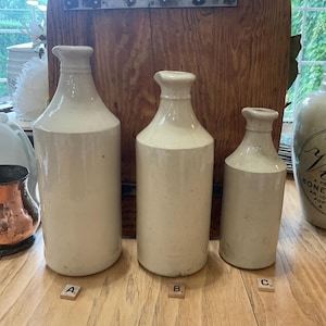 May include: Three antique stoneware bottles of varying sizes, arranged on a wooden surface. The bottles are off-white with a cylindrical shape and narrow necks. The background features a wooden panel and a window.