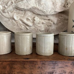 May include: Four cylindrical ceramic pots with a light beige finish and vertical ribbed detailing. Each pot features a smooth rim and a consistent, uniform shape, standing side by side in a row, showcasing a simple, functional design.