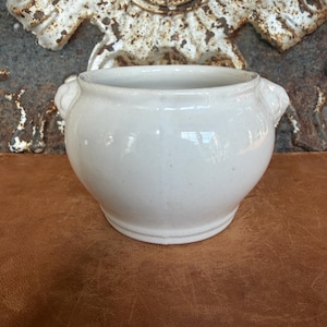 May include: A small, off-white ceramic bowl with a rounded body and a smooth, glossy finish. The bowl features two small, molded handles on opposite sides near the rim, providing a simple, functional design with a clean, uniform appearance.