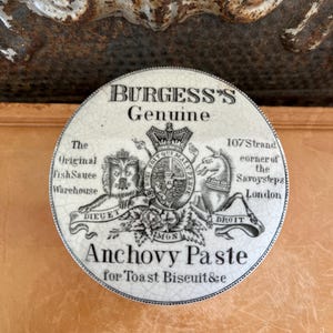 May include: A round, antique ceramic container lid for Burgess's Genuine Anchovy Paste. The lid features a black and white design with text and illustrations, including a lion, unicorn, and a royal crest. The text includes "The Original Fish Sauce Warehouse" and "107 Strand corner of the Savoy steps London."