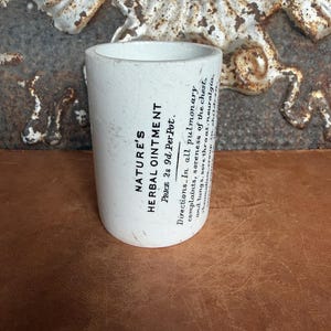 May include: A white cylindrical ceramic pot featuring black printed text that reads NATURE'S HERBAL OINTMENT, Price 2s 9d Per Pot, and Directions: In all pulmonary complaints, soreness of the chest and lungs, sore throat, neuralgia, rheumatism, croup in children.