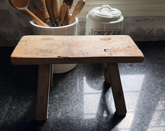 Könnte beinhalten: Ein kleiner, rechteckiger Holzhocker mit vier ausgestellten Beinen und sichtbaren Verbindungen auf der Sitzfläche. Das Holz weist eine natürliche, verwitterte Oberfläche mit verschiedenen Kratzern und Spuren auf Sitz und Beinen auf.