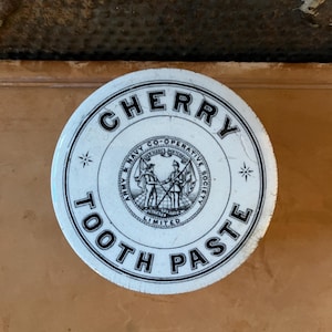May include: A round, white Cherry Tooth Paste tin with black lettering. The center features an illustration of two figures and the words "Army & Navy Co-Operative Society Limited".
