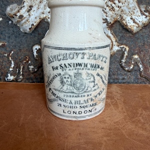 May include: A cream-colored ceramic jar with a cylindrical body and a narrow neck. The front features black printed text reading ANCHOVY PASTE, FOR SANDWICHES &c., BY APPOINTMENT, PURVEYORS TO HER MAJESTY, CROSSE & BLACKWELL, 21 SOHO SQUARE, LONDON.