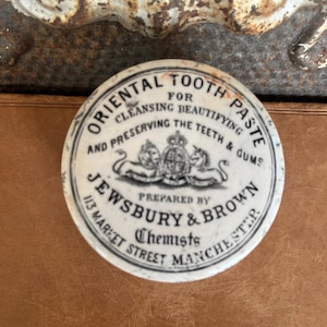 May include: A round white ceramic lid featuring black printed text that reads Oriental Tooth Paste for Cleansing Beautifying and Preserving the Teeth & Gums, prepared by Jewsbury & Brown Chemists, 113 Market Street Manchester, with a royal crest.
