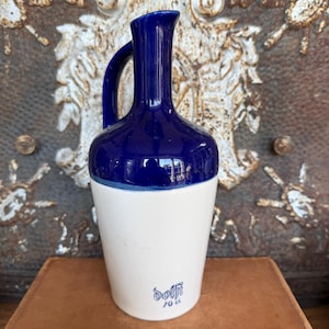 May include: A ceramic pitcher with a glossy dark blue upper section and a matte white lower section. The pitcher features a curved handle and a narrow neck. The text Dolfi and 70 cl are printed in blue on the lower white portion of the vessel.