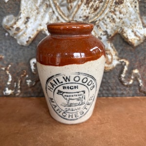 May include: A small, antique cream jar with a brown top and a white base. The jar features the text "HAILWOOD'S RICH CREAM MANCHESTER" in a circular design, with a cow illustration in the center. The jar is likely made of ceramic.