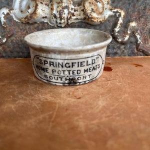 May include: Small, antique stoneware pot with a rounded shape. The pot is off-white with black text that reads "SPRINGFIELD" and "HOME POTTED MEATS SOUTHPORT". The pot has a slightly aged appearance.