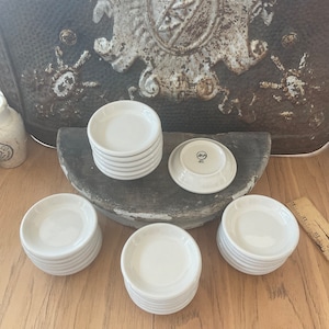 May include: A collection of small, white ceramic saucers. Several stacks of saucers are arranged on a wooden surface, with one saucer flipped over to show the underside. The saucers have a simple, classic design.