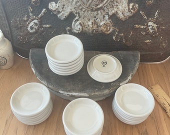 May include: A collection of small, white ceramic saucers. Several stacks of saucers are arranged on a wooden surface, with one saucer flipped over to show the underside. The saucers have a simple, classic design.