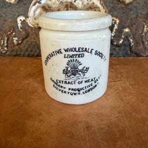 May include: A cylindrical off-white ceramic jar featuring black printed text that reads Cooperative Wholesale Society Limited, Extract of Meat, Grocery Productive Dept, Silvertown, London, with a central graphic of wheat and tools.