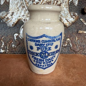May include: A beige ceramic jar with a narrow neck and a flared rim. The front features blue printed text that reads GARRICKS CUMBERLAND DAIRY CO LTD PURE THICK CREAM FRESH DAILY LOW ROW ASPATRIA, centered around a circular emblem with a central symbol.