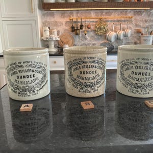 May include: Three cylindrical ceramic marmalade jars with off-white glazes and black printed text reading James Keiller & Sons Dundee Marmalade, Grand Medal of Merit Vienna 1873, and London 1862. Each jar is labeled with a small wooden letter tile A, B, or C.
