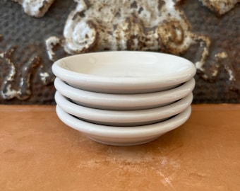 May include: A stack of four small, round, white ceramic dishes with smooth, curved edges. The dishes are uniform in size and color, featuring a glossy finish that reflects light. Each dish has a shallow basin and a simple, clean design.
