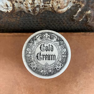 May include: A round, antique ceramic jar lid with the words "Cold Cream" in ornate black lettering. The lid is white with a decorative border featuring floral and scrollwork designs. The background is a warm, copper-toned surface.