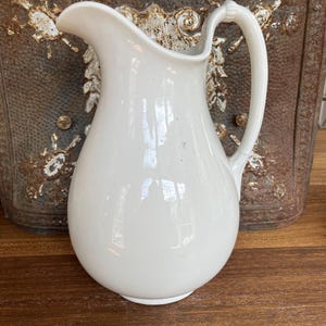 May include: A white ceramic pitcher with a curved spout and a handle. The pitcher has a smooth, glossy finish and a rounded body. The handle is attached at the top and bottom of the pitcher. The background is a rustic metal panel and a wooden surface.