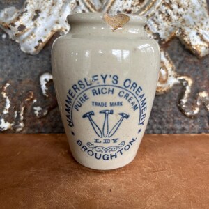 May include: A small, off-white ceramic jar with a blue logo that reads "HAMMERSLEY'S CREAMERY PURE RICH CREAM TRADE MARK LEY BROUGHTON." The jar has a slightly chipped rim and a rounded body. The logo features three hammers.
