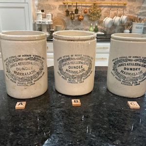 May include: Three off-white ceramic marmalade jars with cylindrical bodies. Each jar features black printed text reading Grand Medal of Merit Vienna 1873, James Keiller & Sons, Dundee Marmalade, and Only Prize Medal for Marmalade London 1862.