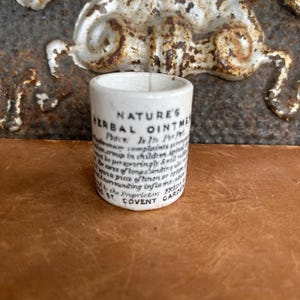 May include: A small, cylindrical white ceramic container with the text "NATURE'S HERBAL OINTMENT" printed on the side. The container has a slightly aged appearance, with visible cracks. The text includes the address "COVENT GARDEN".