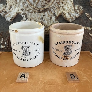 May include: Two antique ceramic Bloater Paste jars from J. Sainsbury's. One is white, the other is tan. Both jars have the text "FRESHLY MADE BLOATER PASTE" and the J. Sainsbury's logo in black. The jars are on a wooden surface.