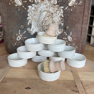 May include: A stack of 10 white ceramic bowls with a wooden handled brush on top. The bowls are stacked in a pyramid shape. The bowls are all the same size and shape. The brush is made of natural bristles and has a wooden handle.