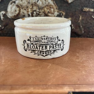 May include: A small, off-white ceramic jar with a slightly cracked rim. The jar features black text that reads "J. Sainsbury's Superior Home Made Bloater Paste London". The jar is cylindrical and sits on a brown surface.