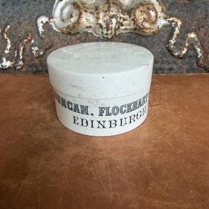 May include: A round white container with a lid, featuring black printed text that reads DUNCAN, FLOCKHART & CO. EDINBURGH. The container has a weathered, matte finish with visible signs of age and wear on the surface and edges of the lid.