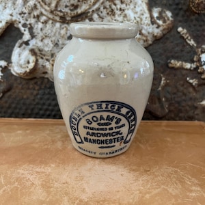 May include: Small, off-white ceramic jar with a rounded body and a short neck. The jar features a blue and white label with text that reads "BOAM'S THICK CREAM, ARDWICK, MANCHESTER". The jar is likely vintage.