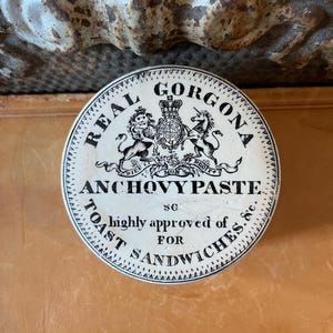May include: A round, vintage tin of Real Gorgona Anchovy Paste, with black text and a crest design on a white background. The text reads "highly approved of for toast sandwiches." The tin is on a brown surface.