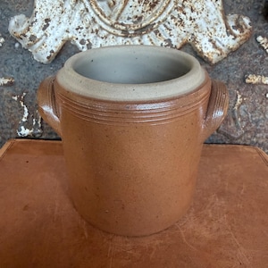 May include: A cylindrical brown stoneware crock featuring two side handles and a smooth, light-colored interior rim. The exterior surface has a speckled texture with horizontal grooved lines encircling the upper portion of the vessel body.
