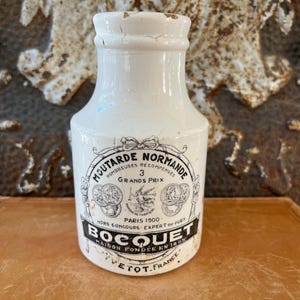 May include: A white ceramic mustard jar with black text and graphics. The jar has a slightly tapered shape and a small opening. The label reads "Moutarde Normande" and "Bocquet, Maison Fondée en 1855, Vetot, France."
