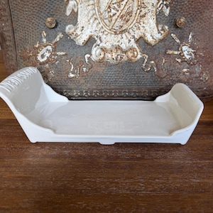 May include: A rectangular white ceramic tray with raised sides and curved ends. The left side features embossed text that reads "WEDGWOOD". The tray has a smooth, glossy finish and a flat base, designed with a shallow interior for holding small items.