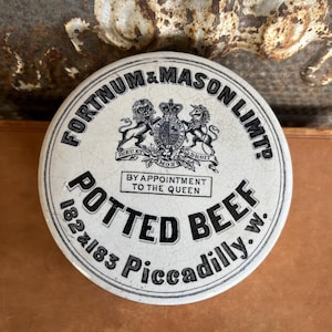 May include: A round, white ceramic lid with black text and a royal crest. The lid reads "FORTNUM & MASON LIMTD" and "POTTED BEEF 182x183 Piccadilly.w." with "BY APPOINTMENT TO THE QUEEN" in the center.