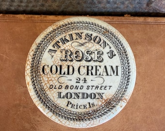 Peut inclure: Un couvercle rond en céramique blanc cassé avec une finition craquelée, portant l'inscription noire : ATKINSON'S ROSE COLD CREAM 24 OLD BOND STREET LONDON PRICE 1S. Le texte est entouré d'une bordure circulaire décorative noire.