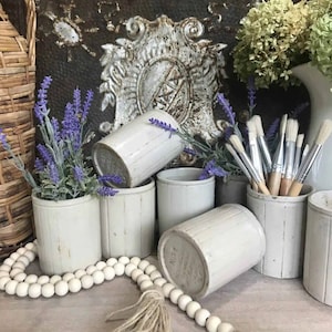 May include: Six white ceramic containers with a ribbed design, arranged on a wooden surface. The containers are filled with paintbrushes and lavender sprigs. A string of white beads with a tassel hangs in the foreground.