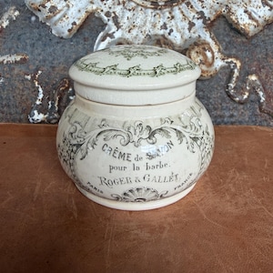 May include: A cream-colored ceramic jar with a matching lid, featuring dark green decorative scrollwork and text that reads Crème de Savon pour la barbe, Roger & Gallet, Paris, France. The container has a rounded body and a smooth finish.