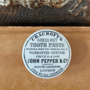 May include: A vintage, round, white and black tin of Cracroft's Areca Nut Tooth Paste. The text on the tin reads "Prepared from the Cincalese Recipe" and "John Pepper & Co. Limited, Bedford Laboratory, London. Sold by all Chemists."