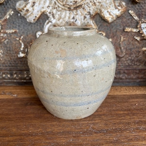May include: A small, round, ceramic vase with a light beige base and horizontal blue stripes. The vase has a slightly textured surface and a small opening at the top. The background features a decorative metal element.