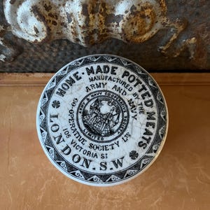 May include: A round, antique ceramic jar lid with black and white text and a central emblem. The text reads "HOME-MADE POTTED MEATS" and "ARMY & NAVY CO-OPERATIVE SOCIETY LTD." The lid is cracked, showing its age.