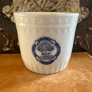 May include: A white ceramic crock with a blue emblem featuring a tree and the text "Mirlands Co. Confitures Fruits". The crock has a ridged rim and a slightly flared shape. It sits on a brown surface.