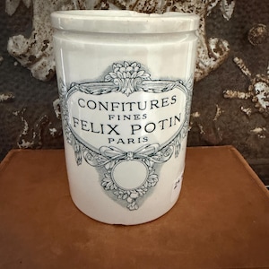 May include: A white ceramic cylindrical jar featuring a dark green decorative label with the text CONFITURES FINES FELIX POTIN PARIS. The label includes an ornate border with floral and ribbon motifs, centered on the front of the smooth, glossy vessel.