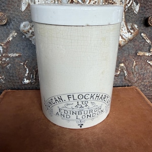 May include: A cylindrical cream-colored ceramic storage jar with a matching lid. The jar features a fine crackle glaze finish and black printed text that reads DUNCAN, FLOCKHART & CO LTD EDINBURGH AND LONDON with decorative scrollwork.