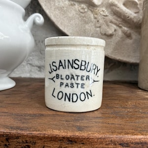 May include: An off-white ceramic pot with the text "J. SAINSBURY BLOATER PASTE LONDON" in black. The pot is cylindrical with a small lid. Other ceramic items and a wooden surface are in the background.