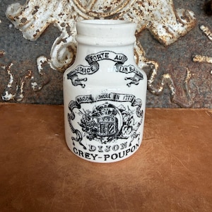 May include: A white ceramic Grey-Poupon mustard jar featuring black printed text and graphics. The label includes the text "Moutarde Forte au Vin Blanc," "Maison Fondée en 1777," and "Dijon." The jar has a cylindrical body with a narrow neck and a wide opening.