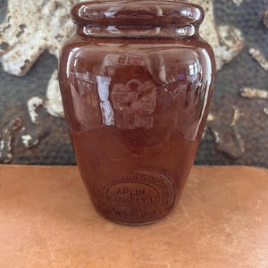 May include: A brown ceramic jar with a rounded body and a lid. The jar has a glossy finish and a circular stamp with text that reads "Western Counties Creamery, Aplin Barrett Ltd, Yeovil, Marston, Westbury."