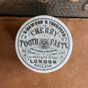 May include: A vintage, round, white and black Cherry Tooth Paste tin from S. Maw, Son & Thompson. The tin features the text "Cherry Tooth Paste" and "For Cleaning & Preserving the Teeth and Gums." The tin is from Aldersgate Street, London, England.