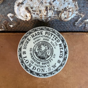 May include: A circular white ceramic lid featuring black printed text that reads Home-Made Potted Meats, Manufactured by the Army and Navy Co-operative Society Limited, 105 Victoria St, London, S.W, with a central emblem of two figures.