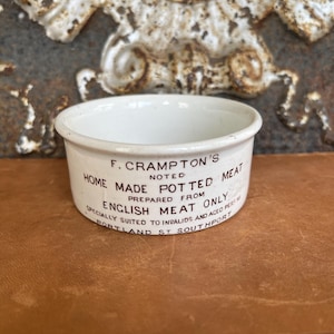May include: A small, white ceramic pot with a rounded rim. The pot has text in dark brown ink that reads "F. Crampton's Noted Home Made Potted Meat". The text continues with "Prepared from English Meat Only" and "Portland St. Southport".